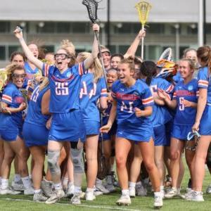 The women's lacrosse team embrace each other and cheer victoriously on the 场. They wear blue Sagehens uniforms with white and orange accent colors.